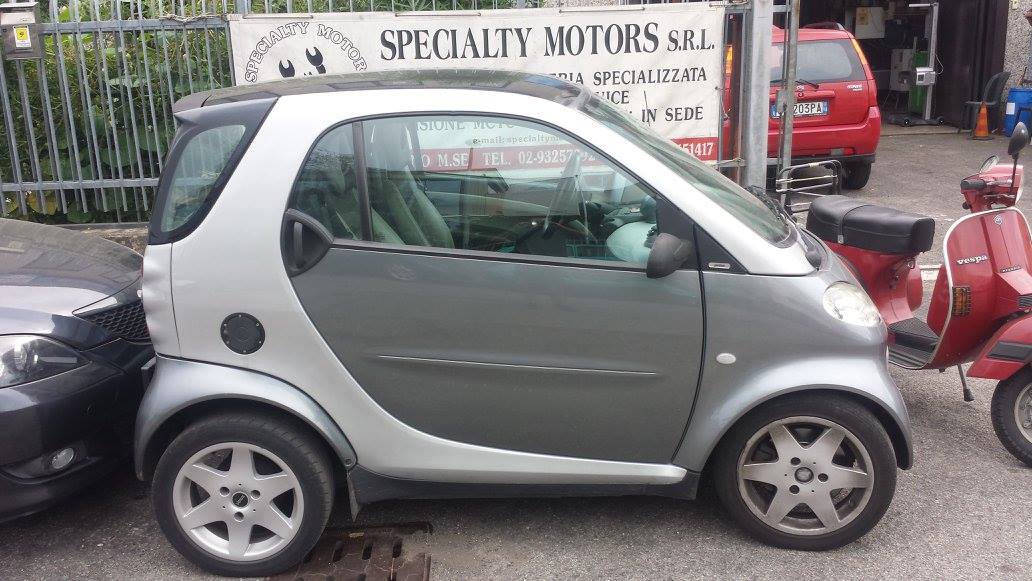 Una Smart argentata parcheggiata vicino a uno scooter rosso e a un cartello commerciale.