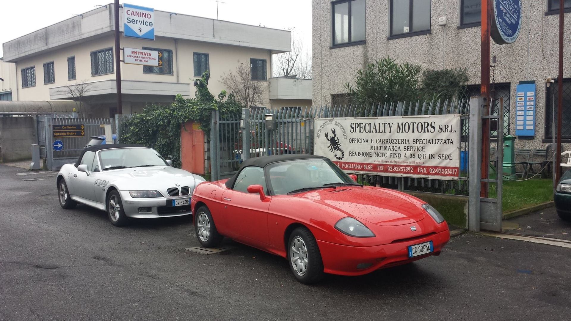 Due decappottabili, una BMW argentata e una Fiat rossa, parcheggiate su una strada di fronte a un'officina.