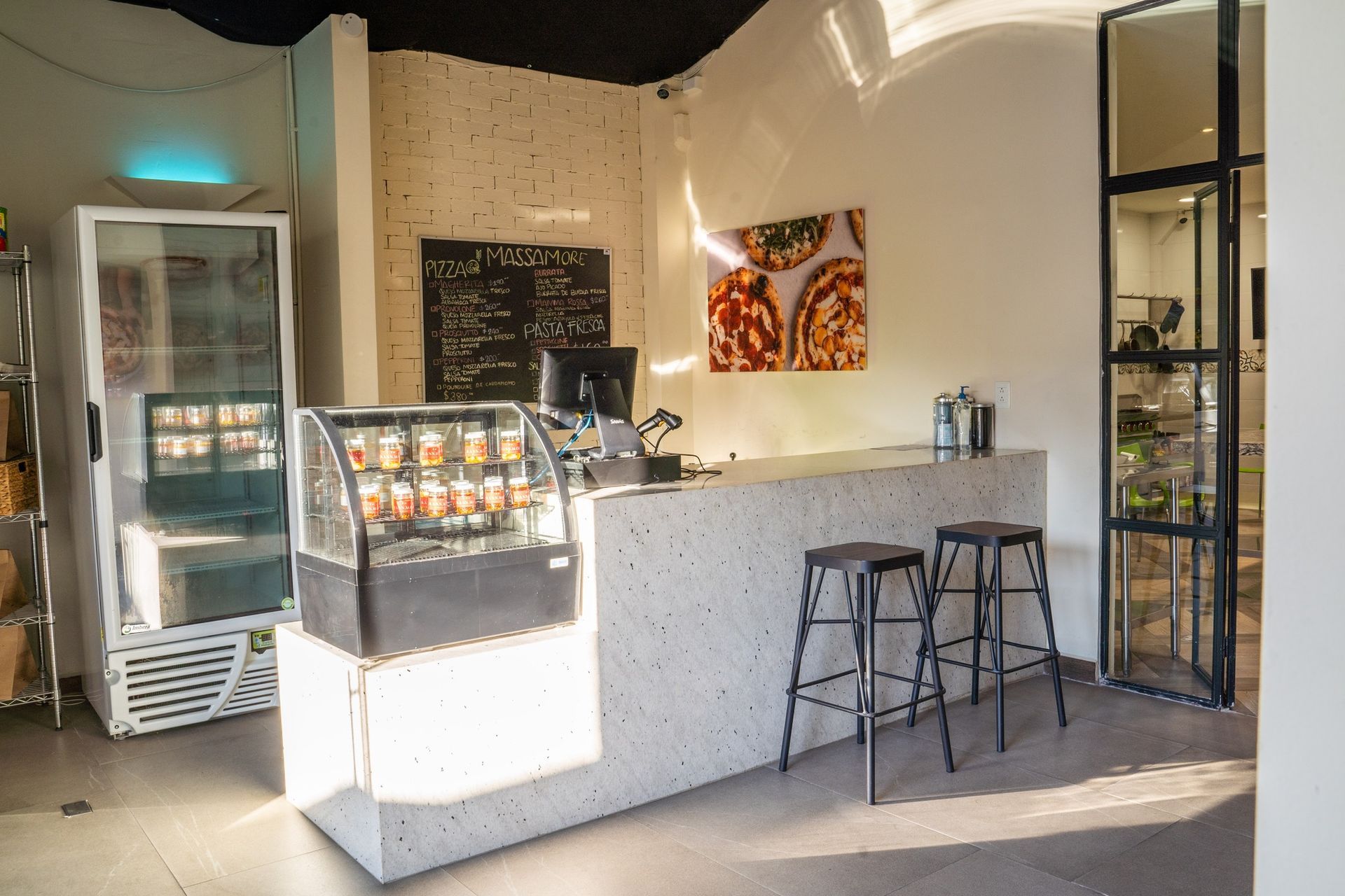 Mostrador de cafetería moderno con vitrina de pasteles, taburetes y refrigerador en un interior luminoso.