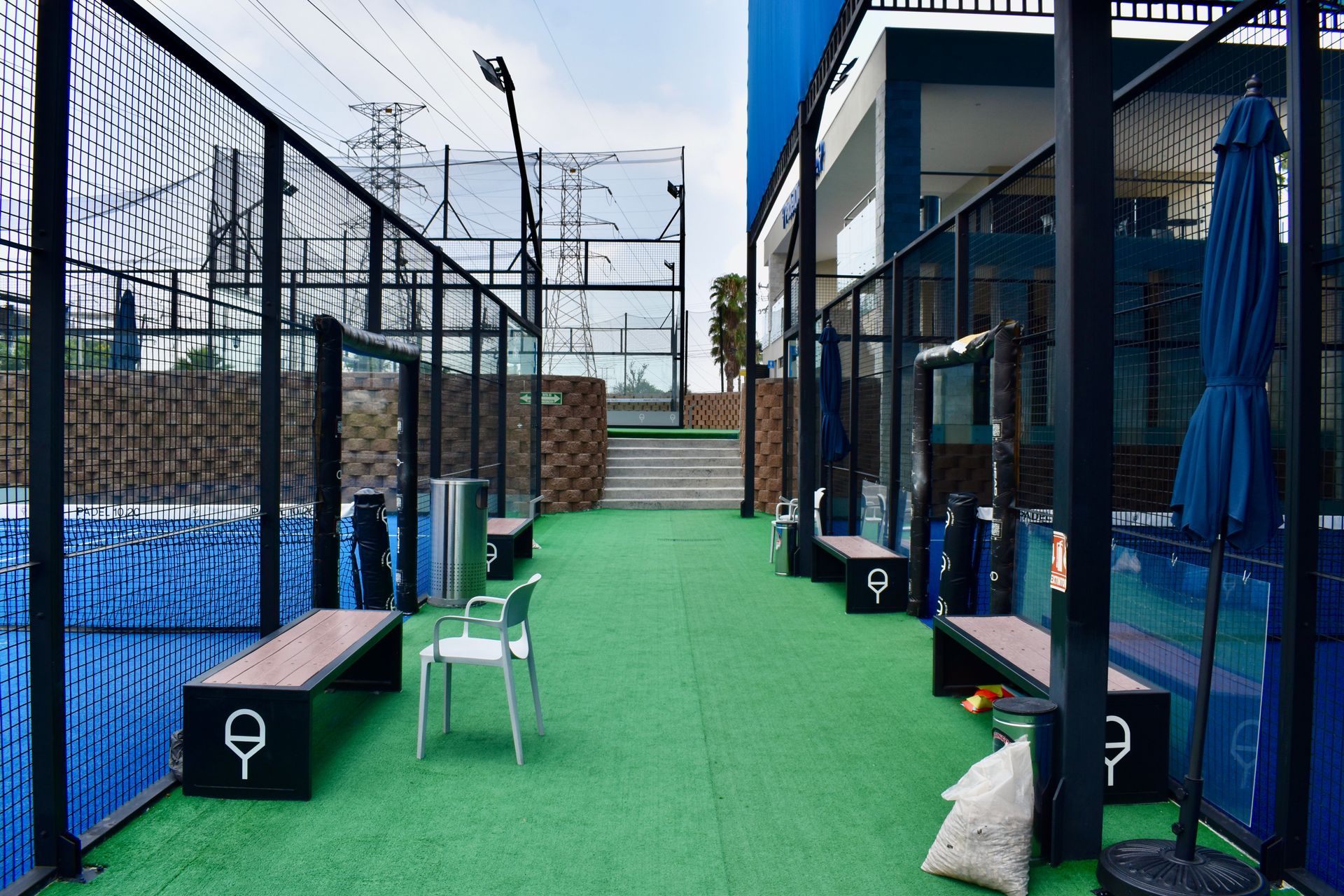 Cancha deportiva al aire libre con césped verde, red azul, bancos y sillas junto a un edificio.