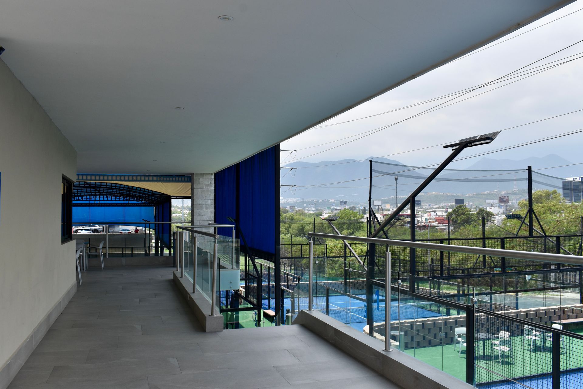 Pasillo cubierto con balcón con vistas a una piscina y a las montañas, con cortinas azules y barandilla de cristal.