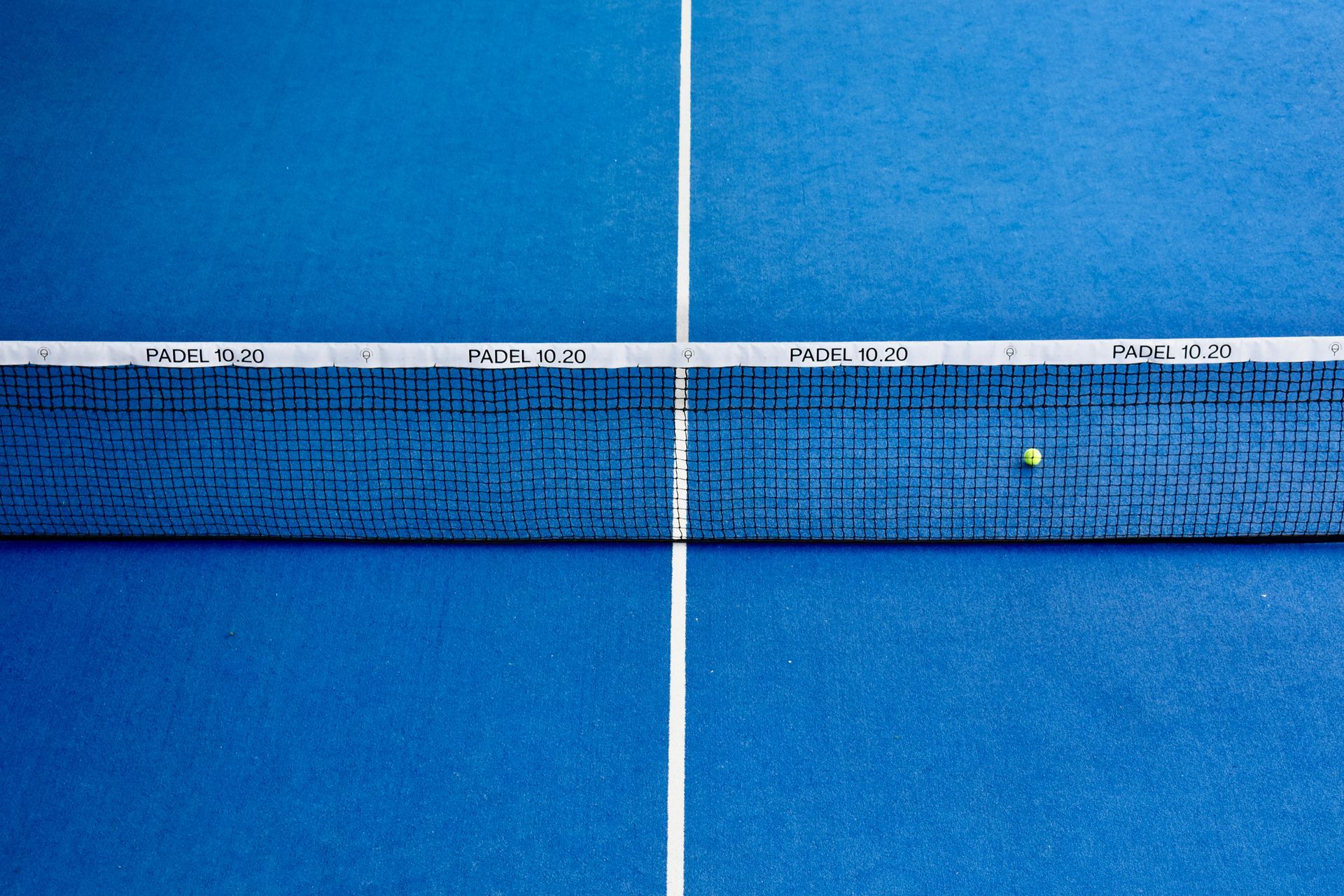 Pista de tenis azul con una red en el centro y una pelota amarilla cerca del centro derecho.