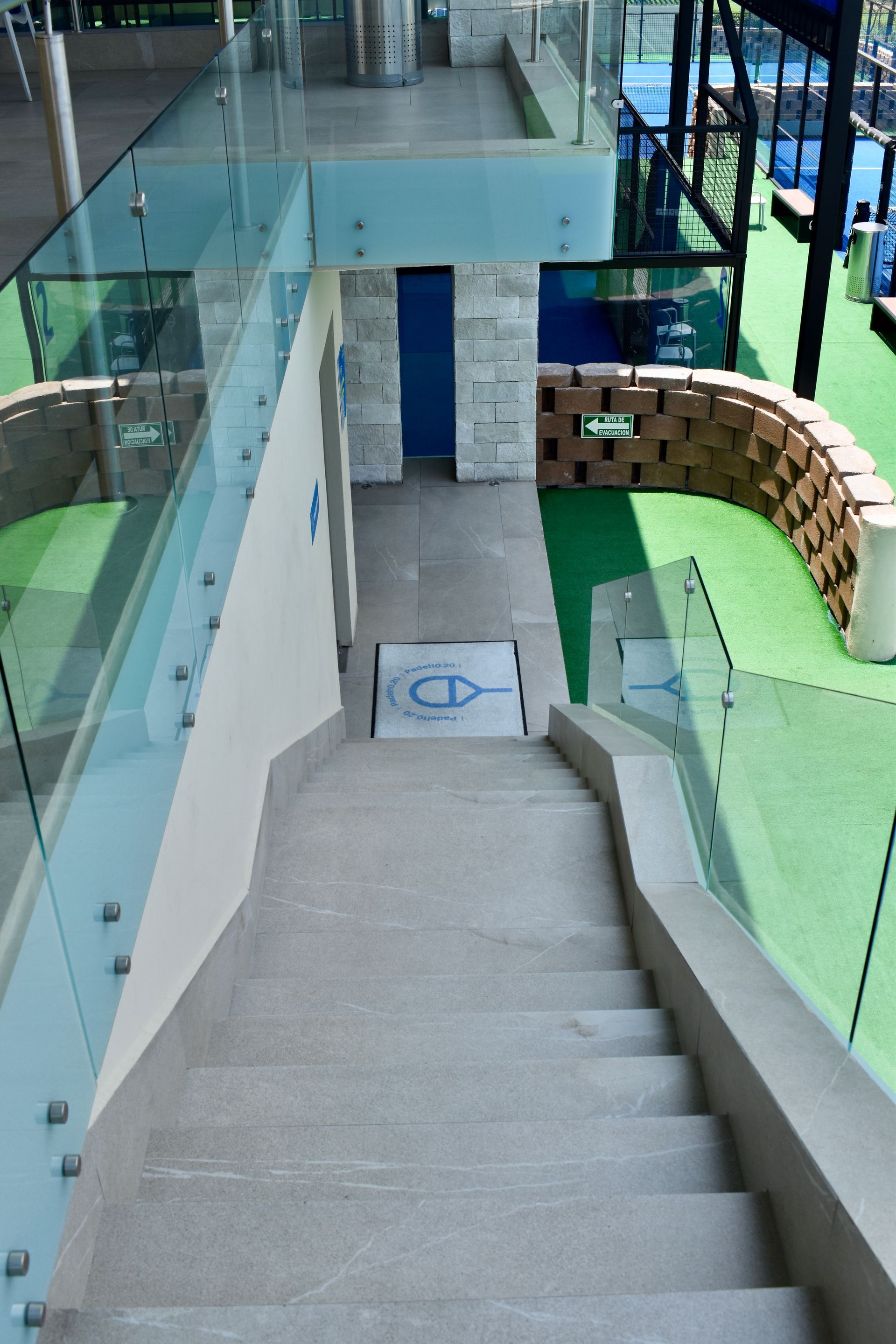 Una empinada escalera de hormigón, junto a barandillas de cristal, conduce a una puerta azul y a una pasarela cubierta.