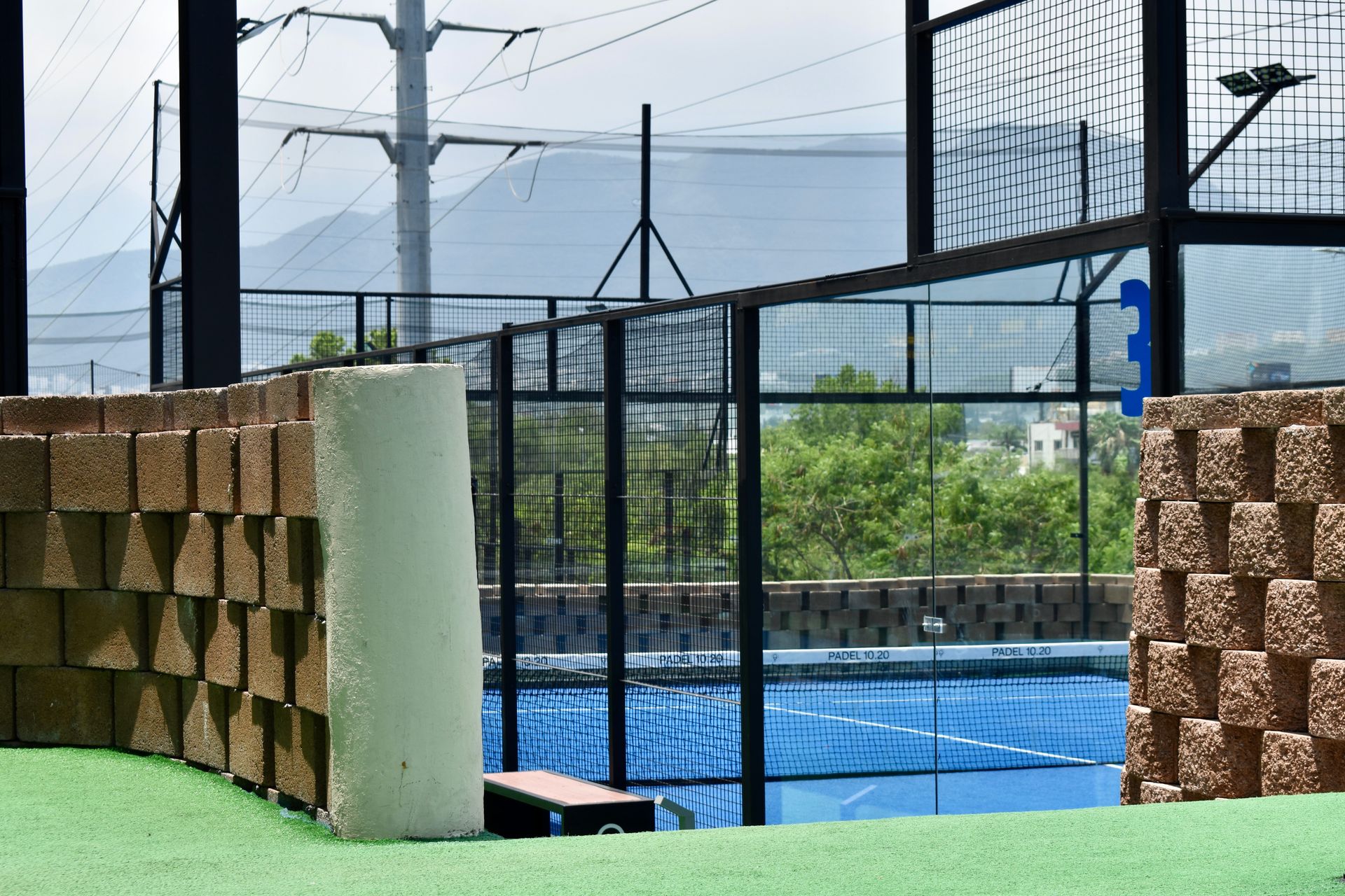 Cancha deportiva al aire libre con superficie azul, césped verde y valla de alambre junto a muros de ladrillo apilados.