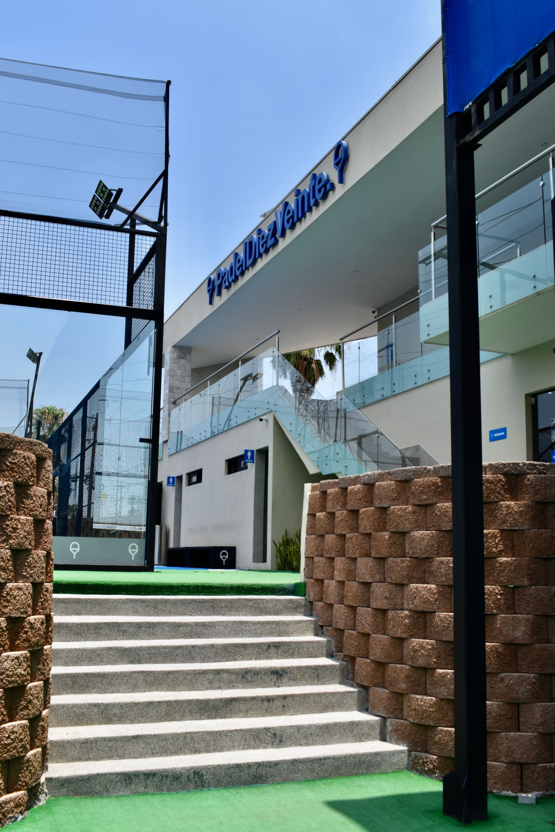 Entrada moderna al edificio con escalones, fachada de cristal y letreros azules bajo un cielo despejado.