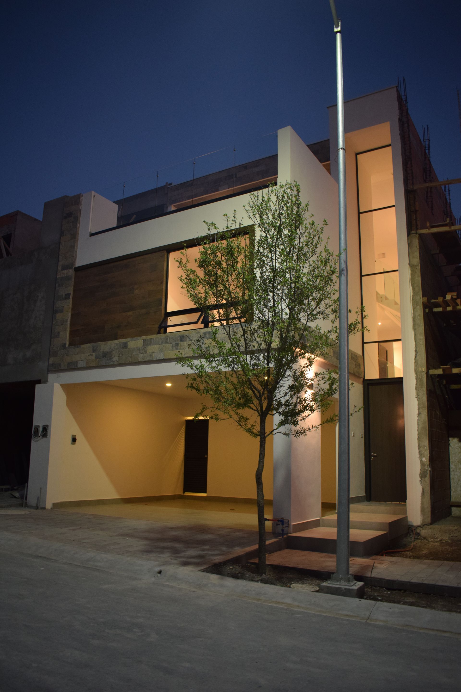 Exterior de una casa moderna de dos plantas al anochecer, con ventanas iluminadas, una cochera y un árbol en la fachada.
