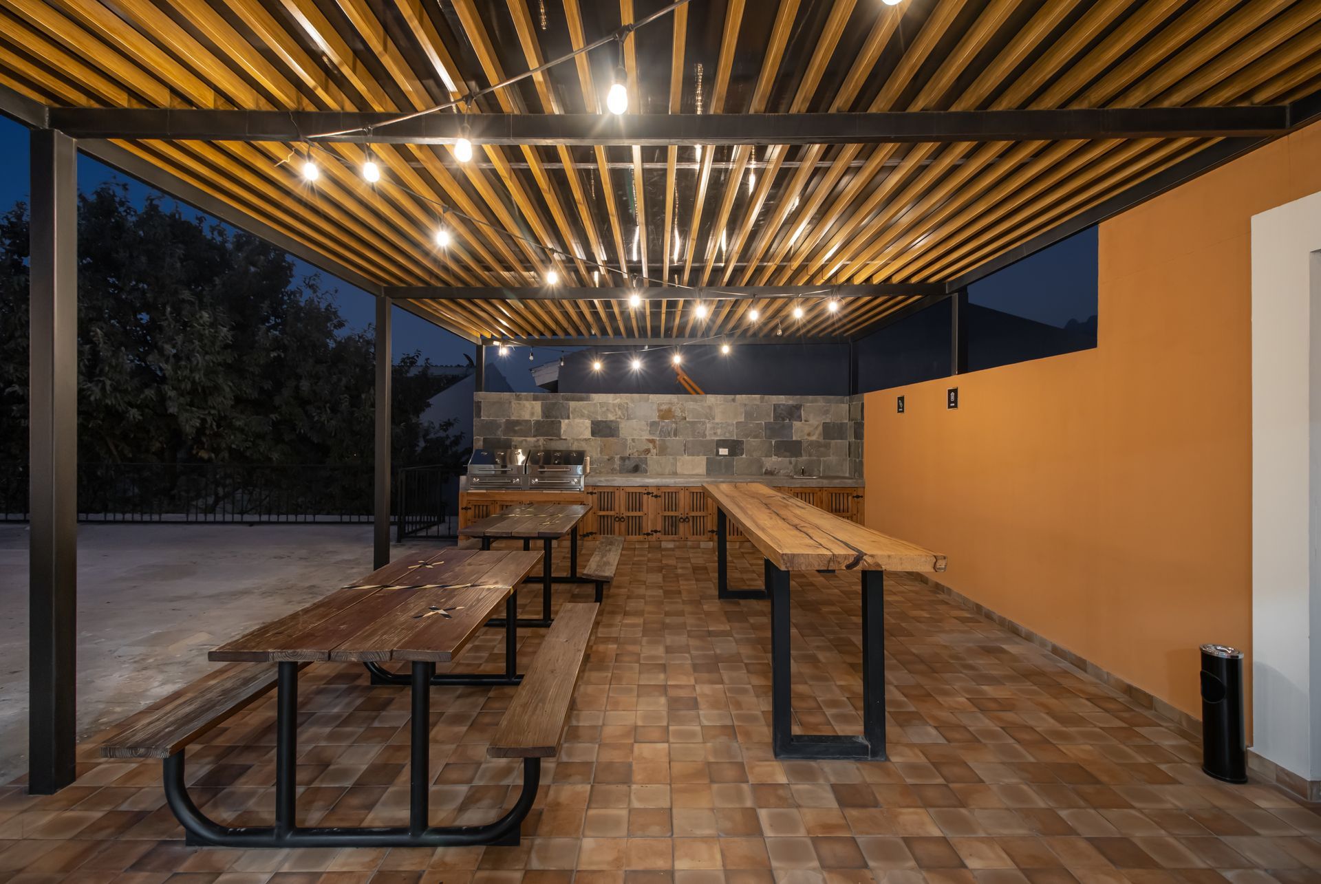 Comedor exterior cubierto con mesas de picnic de madera, guirnaldas de luces y un muro de piedra al atardecer.