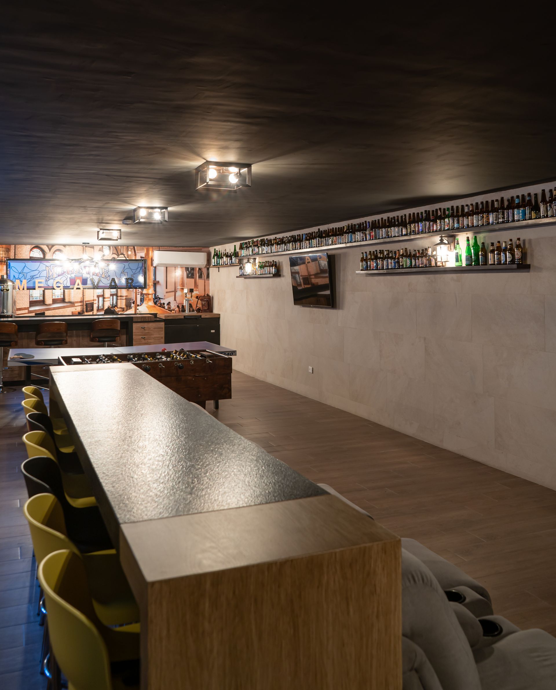 Interior de bar moderno con una larga barra de madera, taburetes verdes y estantes de pared llenos de botellas.
