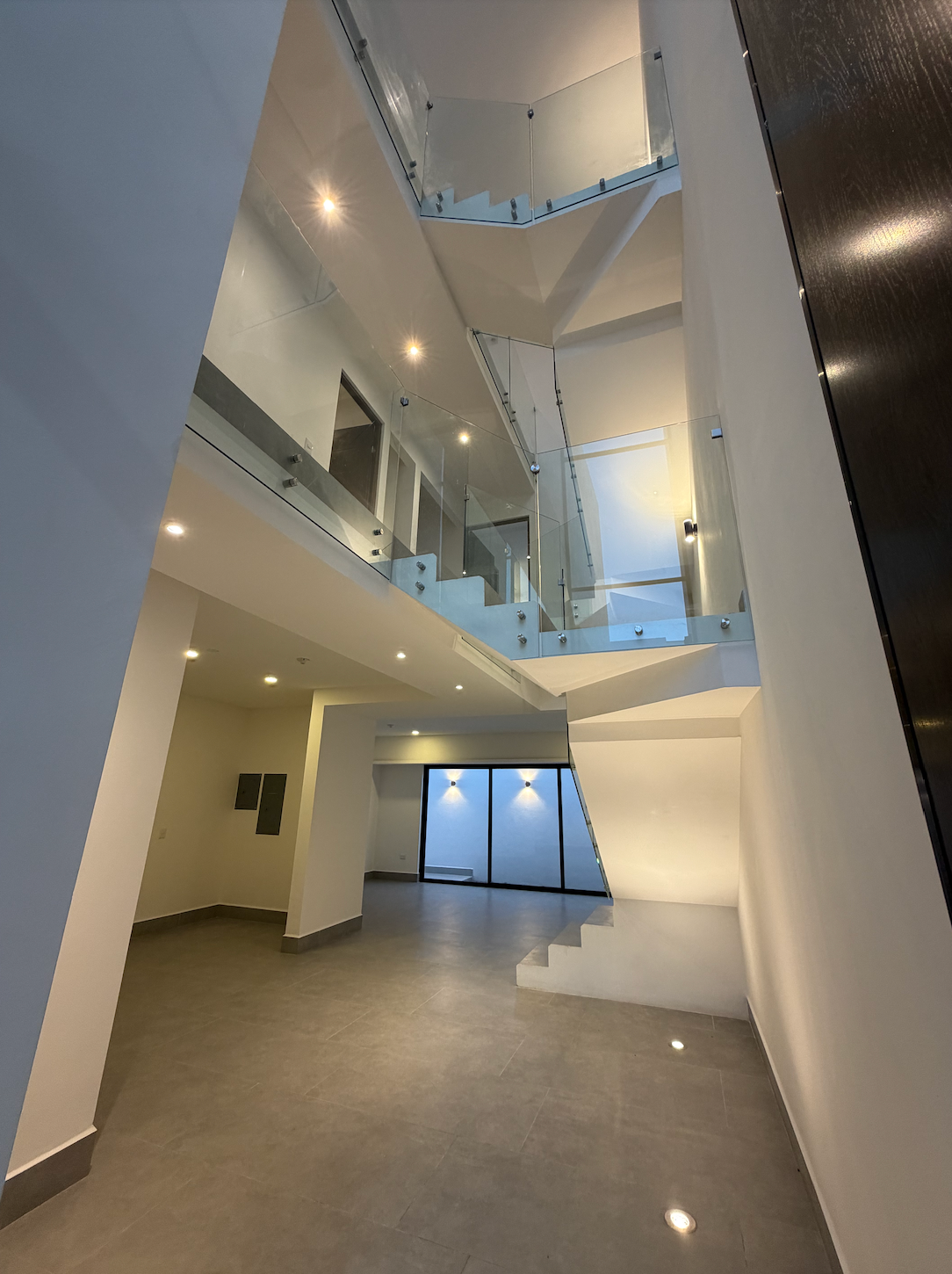 Interior de atrio moderno con balcones curvos, paredes blancas y puertas de cristal bajo luces cálidas en el techo.
