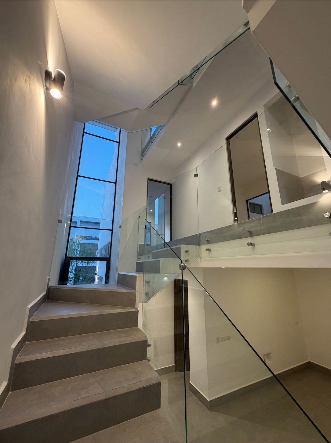 Escalera moderna con barandilla de cristal, escalones alfombrados y grandes ventanales tintados de azul.