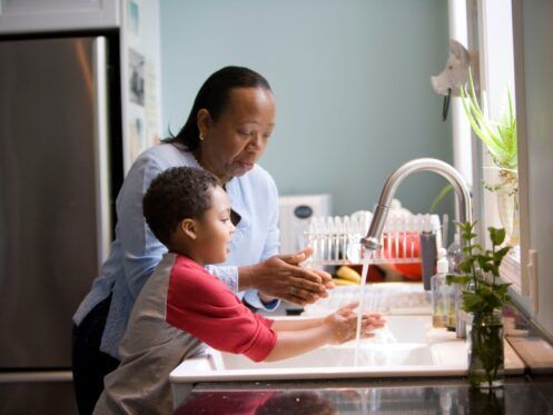 Woman and child washing hands at a kitchen sink. The child is Black and wearing a red and gray shirt.