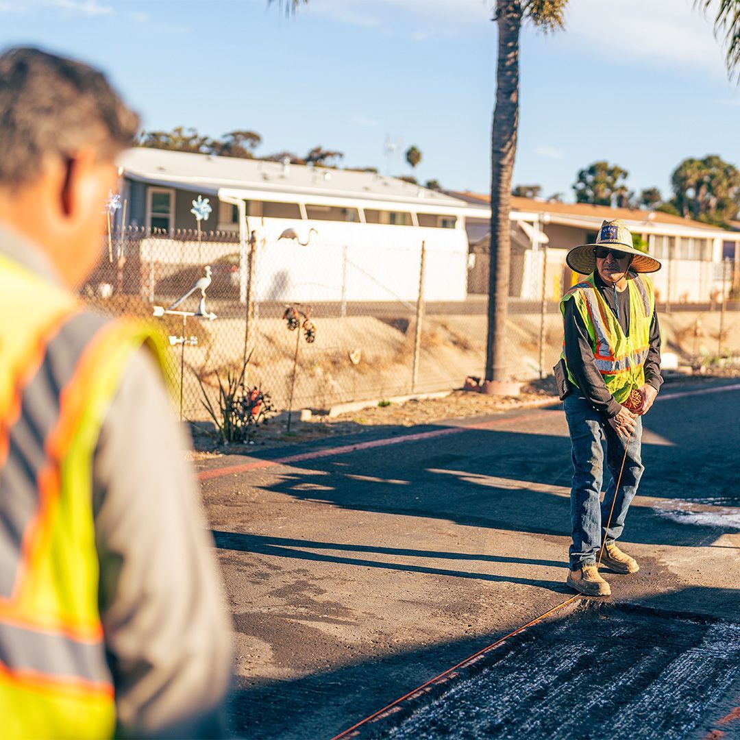 Road workers in reflective vests patching asphalt on a residential street beside a white fence.