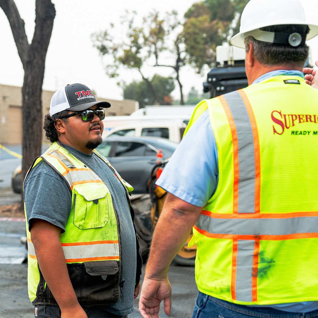 Two utility workers in neon safety vests talk at a roadside work site.