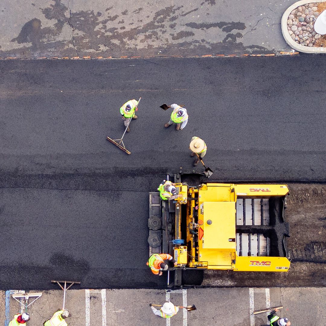 Roadwork crew around a yellow asphalt paver on a paved street, viewed from above.