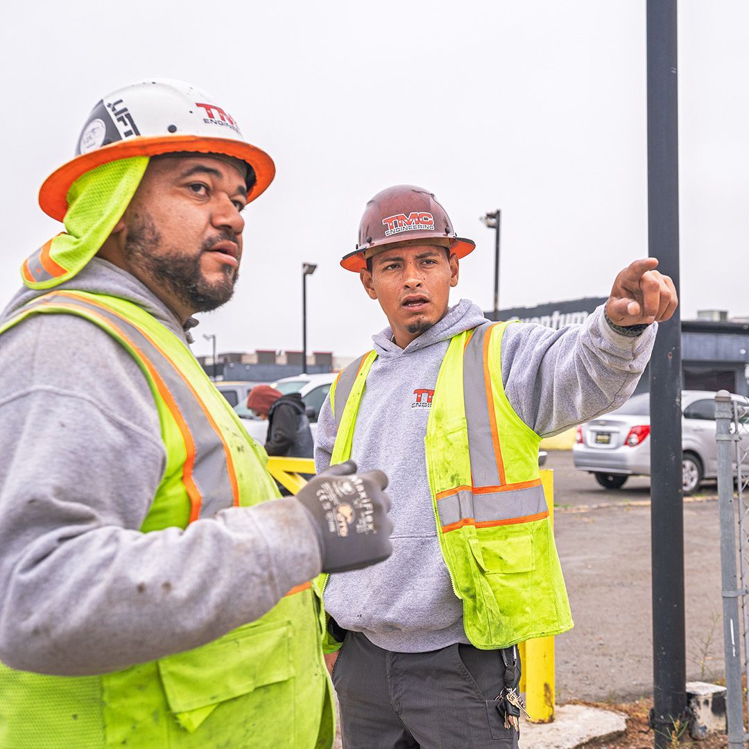 Two construction workers in safety vests and hard hats gesturing at a worksite in a parking lot
