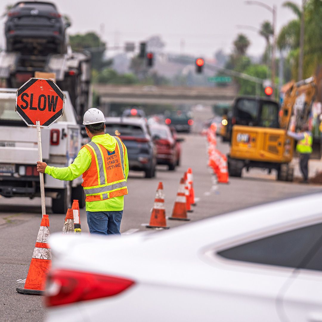 Road construction worker in reflective vest directs traffic near orange cones and a slow sign.