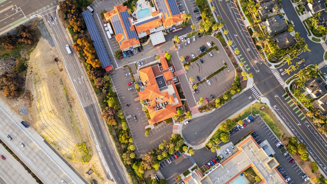 Aerial view of a parking lot beside orange-roofed buildings and a road with cars.