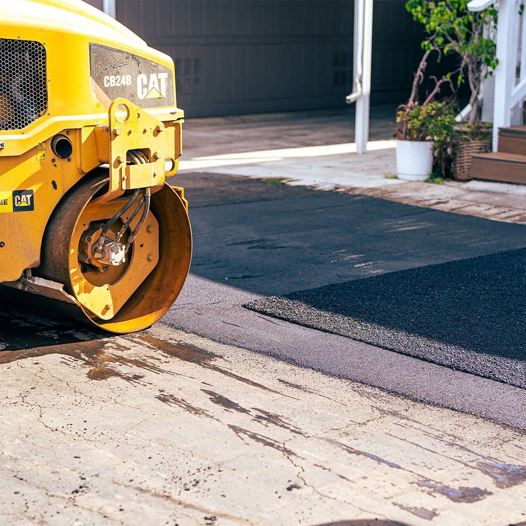 Yellow road roller compacts fresh asphalt beside a sidewalk and building entrance.
