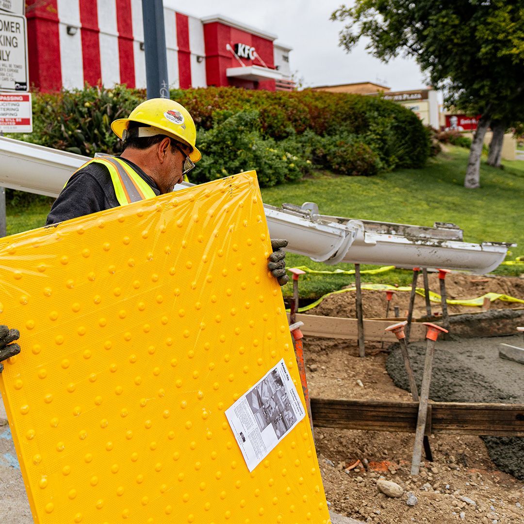 Worker in yellow hard hat and vest carrying a large yellow panel at a roadside construction site.