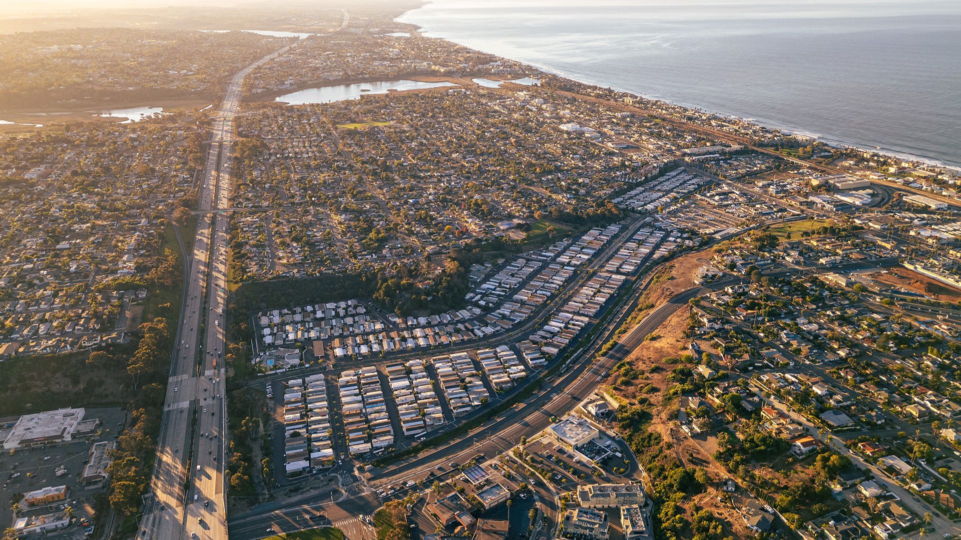 Aerial view of a coastal city at sunset, with dense neighborhoods, roads, and the ocean to the right