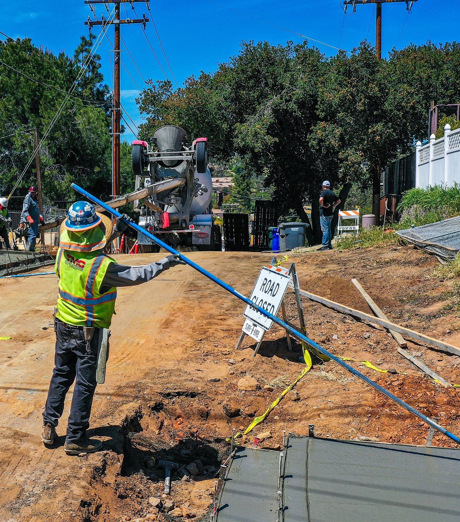 Worker in a neon vest gestures at a muddy roadside construction site with equipment and trees in the background