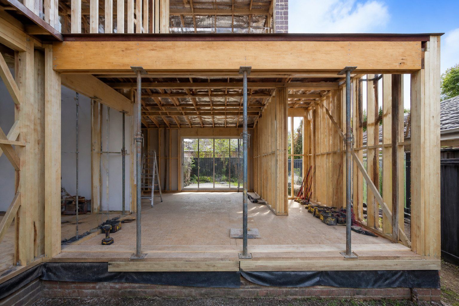 Unfinished house interior with exposed wooden framing and support poles, opening to a backyard.