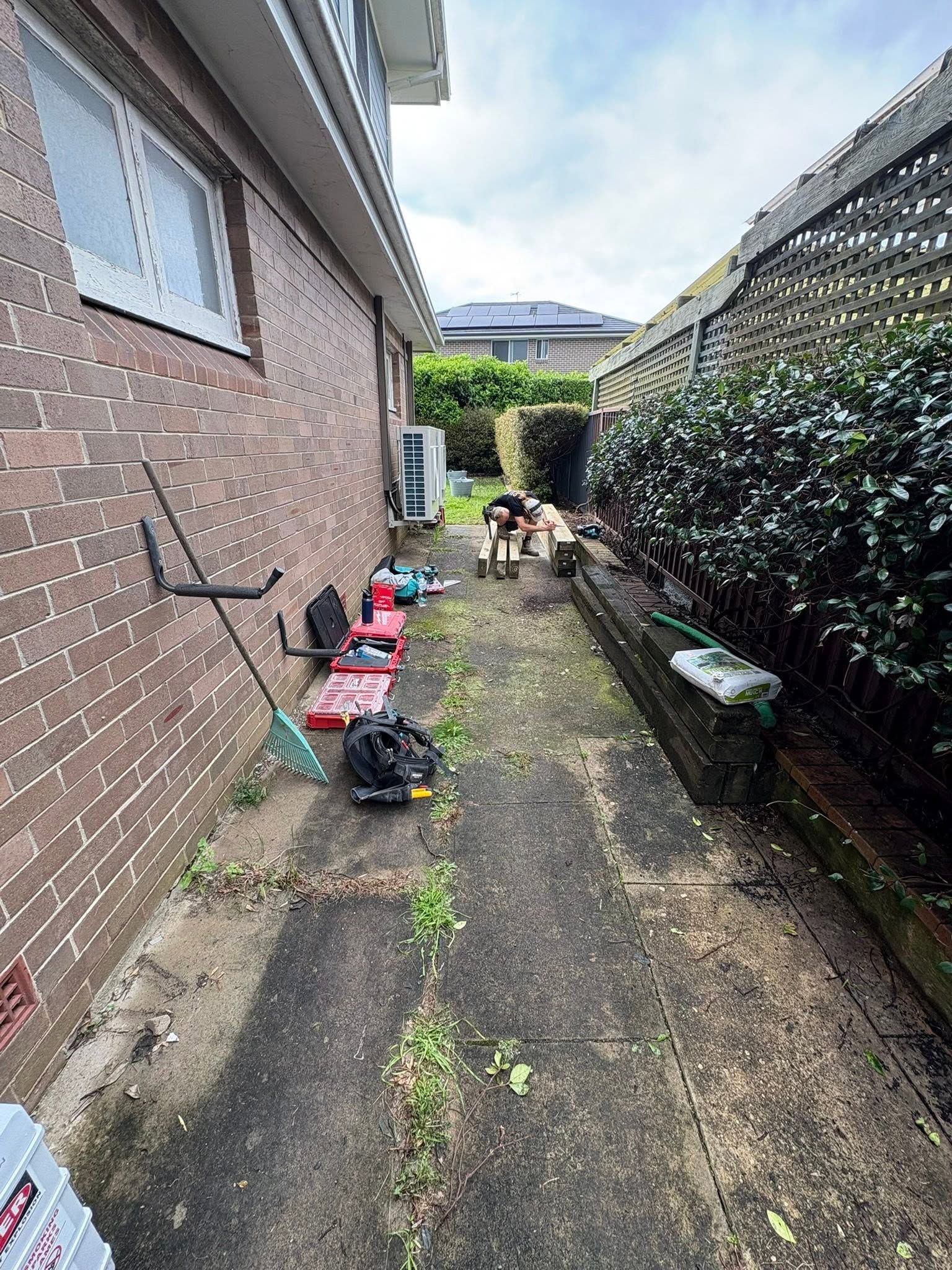 Narrow alley beside a brick house, with tools and clutter on the ground and a dog near the hedge at the far end