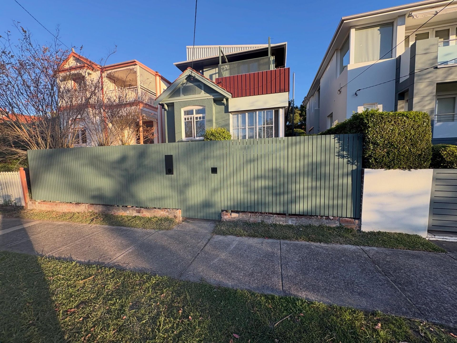 Residential house behind a green fence on a sunny street with a sidewalk and trees