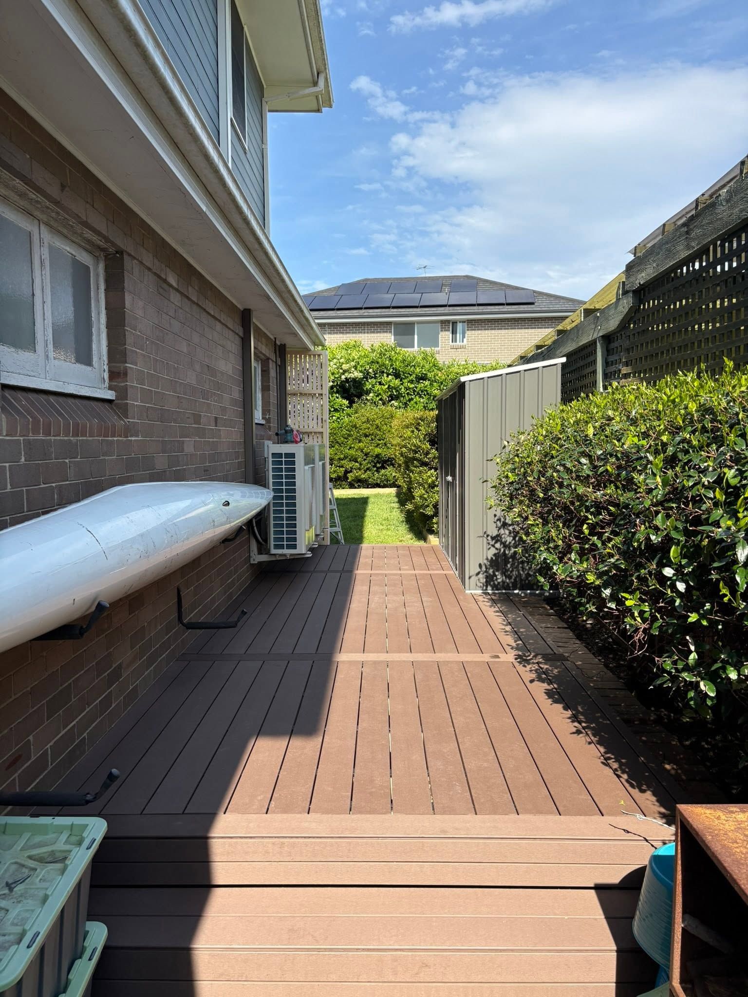 Side yard walkway with wooden deck, brick house wall, hedge, and bright sky.
