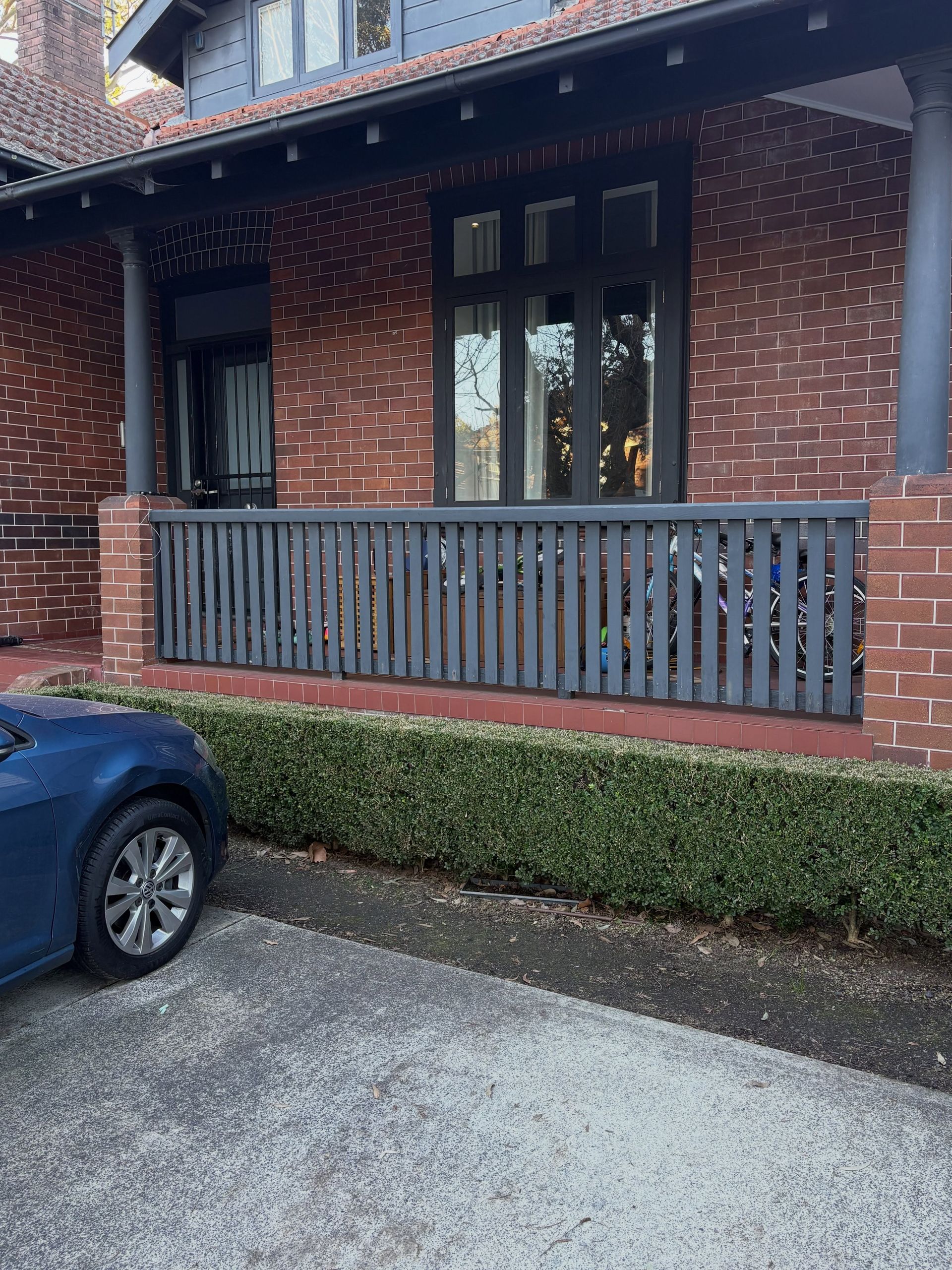 Brick house porch with gray railing, dark window, trimmed hedge, and parked blue car in front