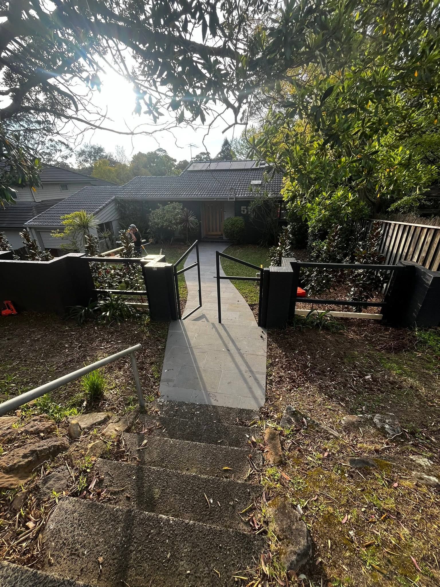 Front path to a house with black gates, bordered by trees and bushes in sunlight