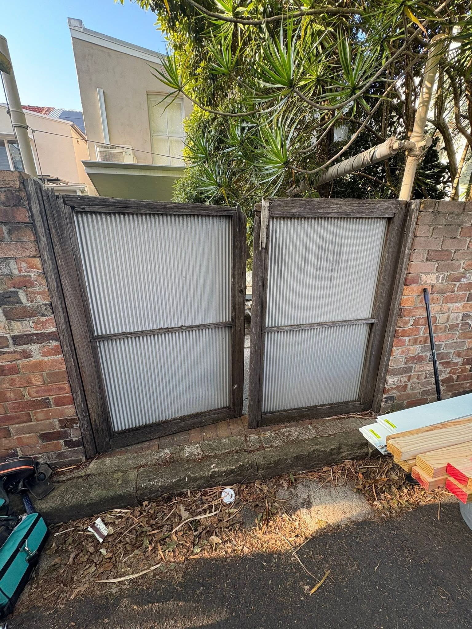 Two corrugated metal gates in a brick wall, with overgrown trees behind and a paved street in front