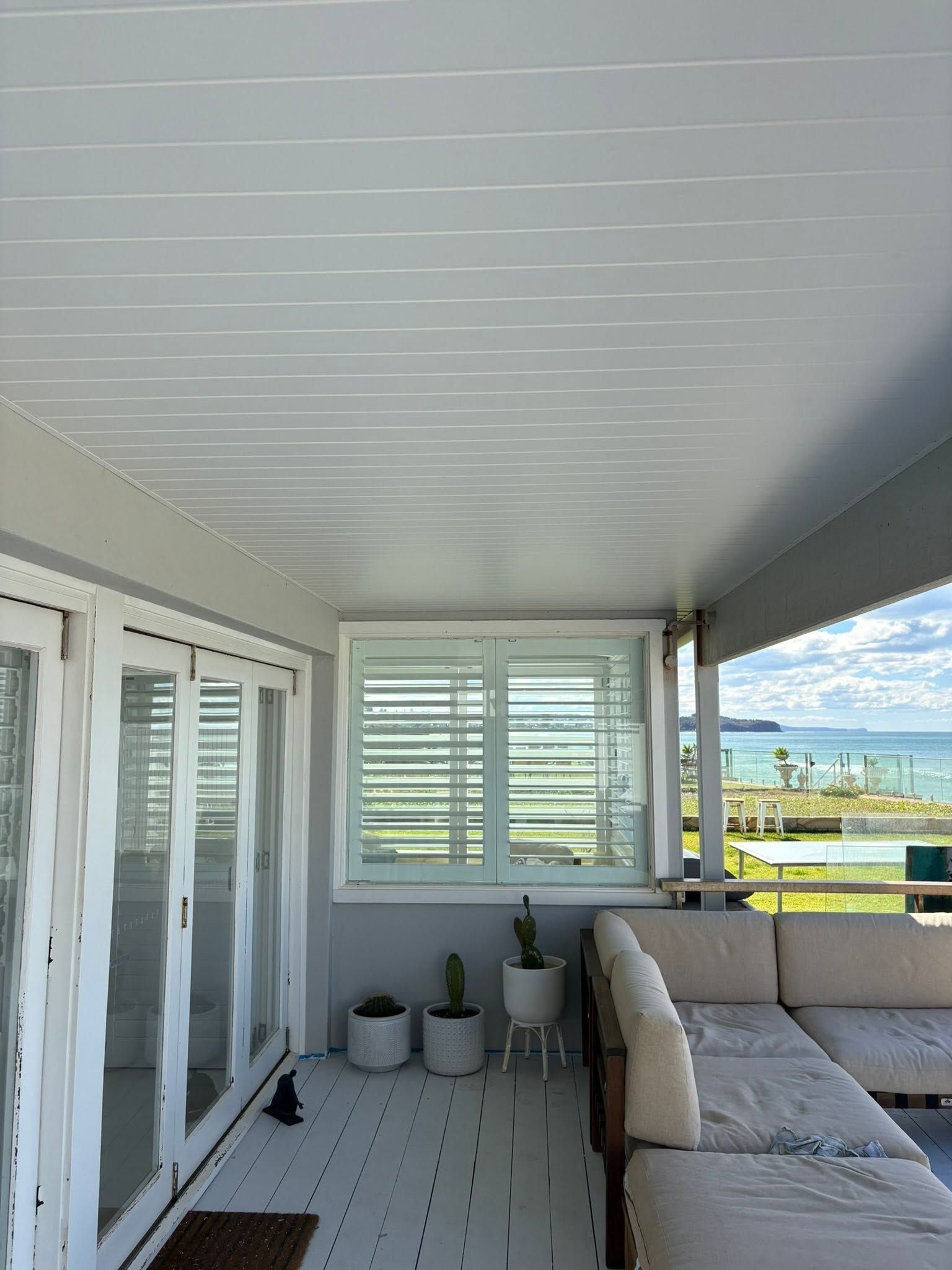 Covered balcony with white shutters, potted plants, and a light gray sofa overlooking a waterfront view