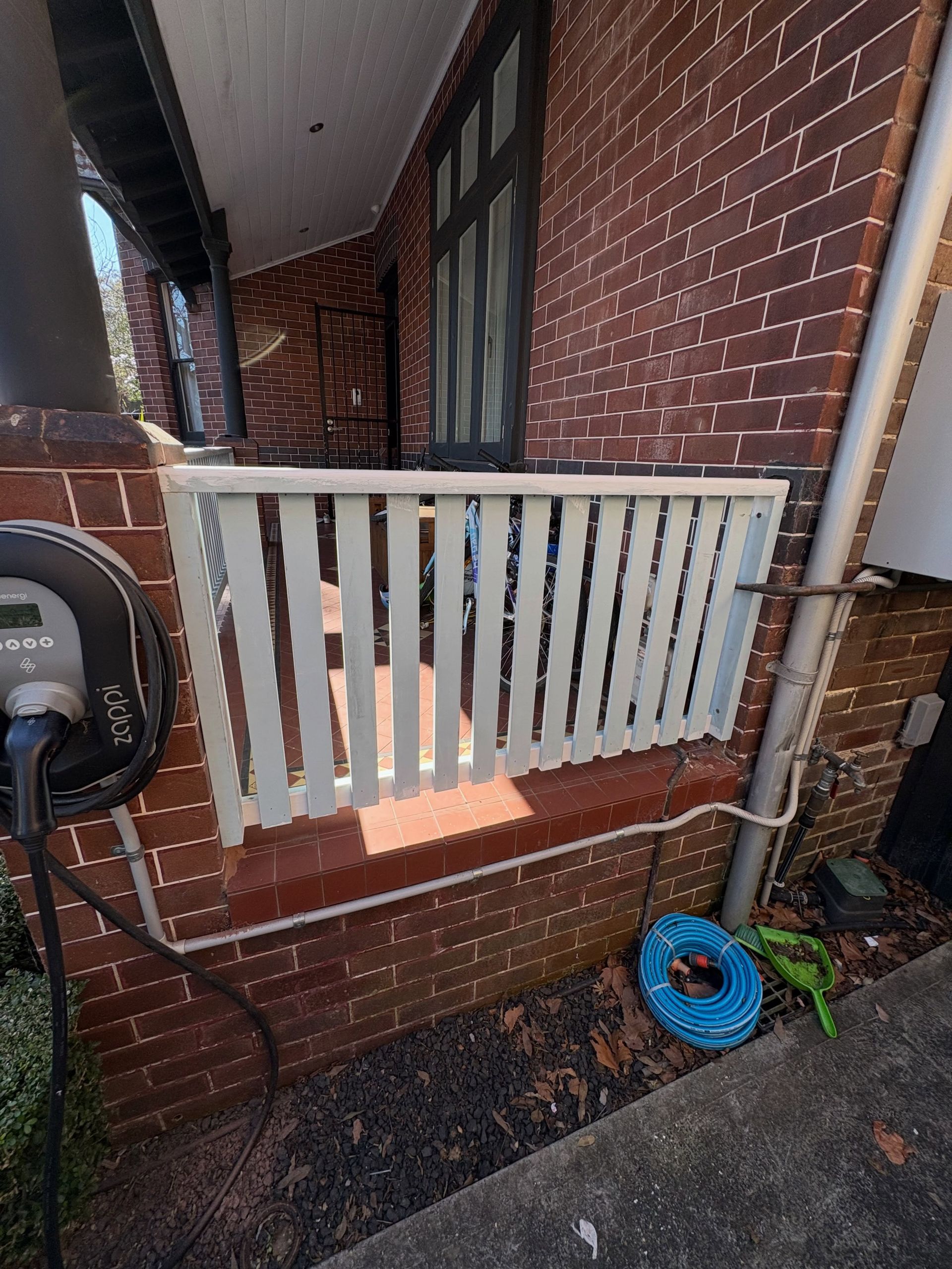 Brick house side porch with white railing, downspout, and coiled blue hose.