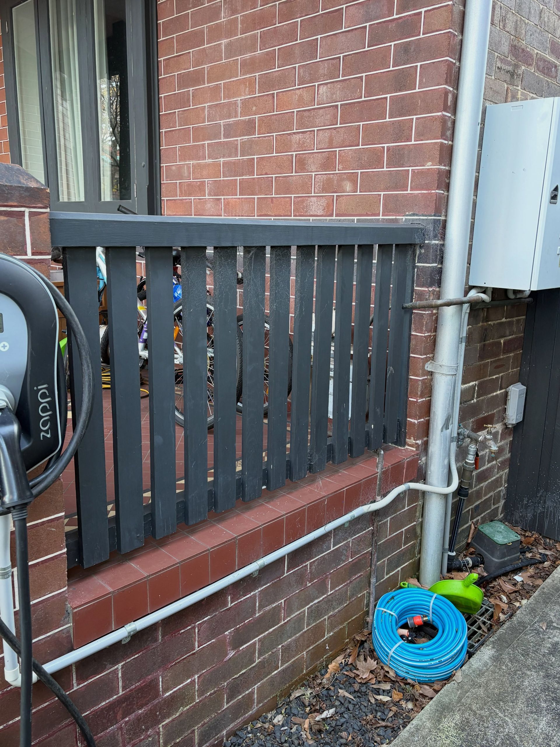 Black wooden railing on a brick building beside an air conditioner and blue hose reel on pavement