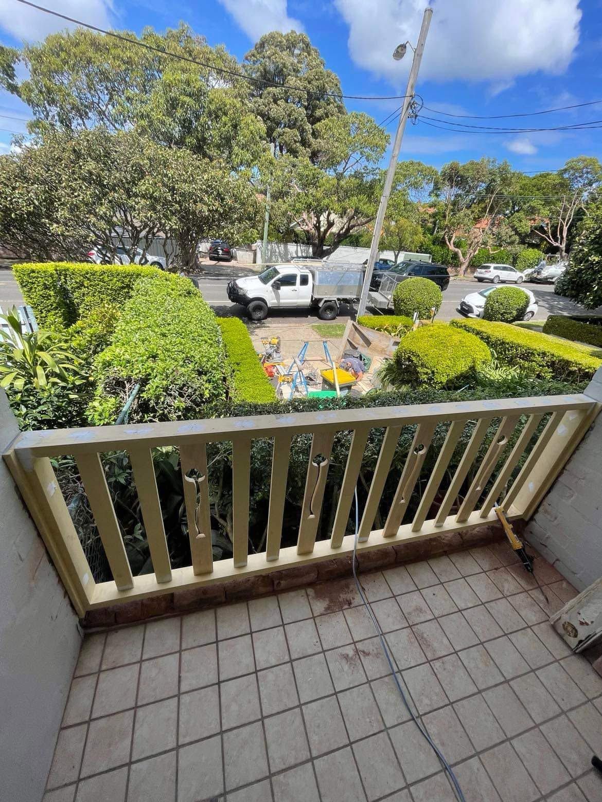 Balcony view of a leafy street with parked cars, garden beds, and a small patio with chairs and table