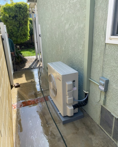 Air conditioning unit mounted on a gray base, next to a pale green wall, outdoors.