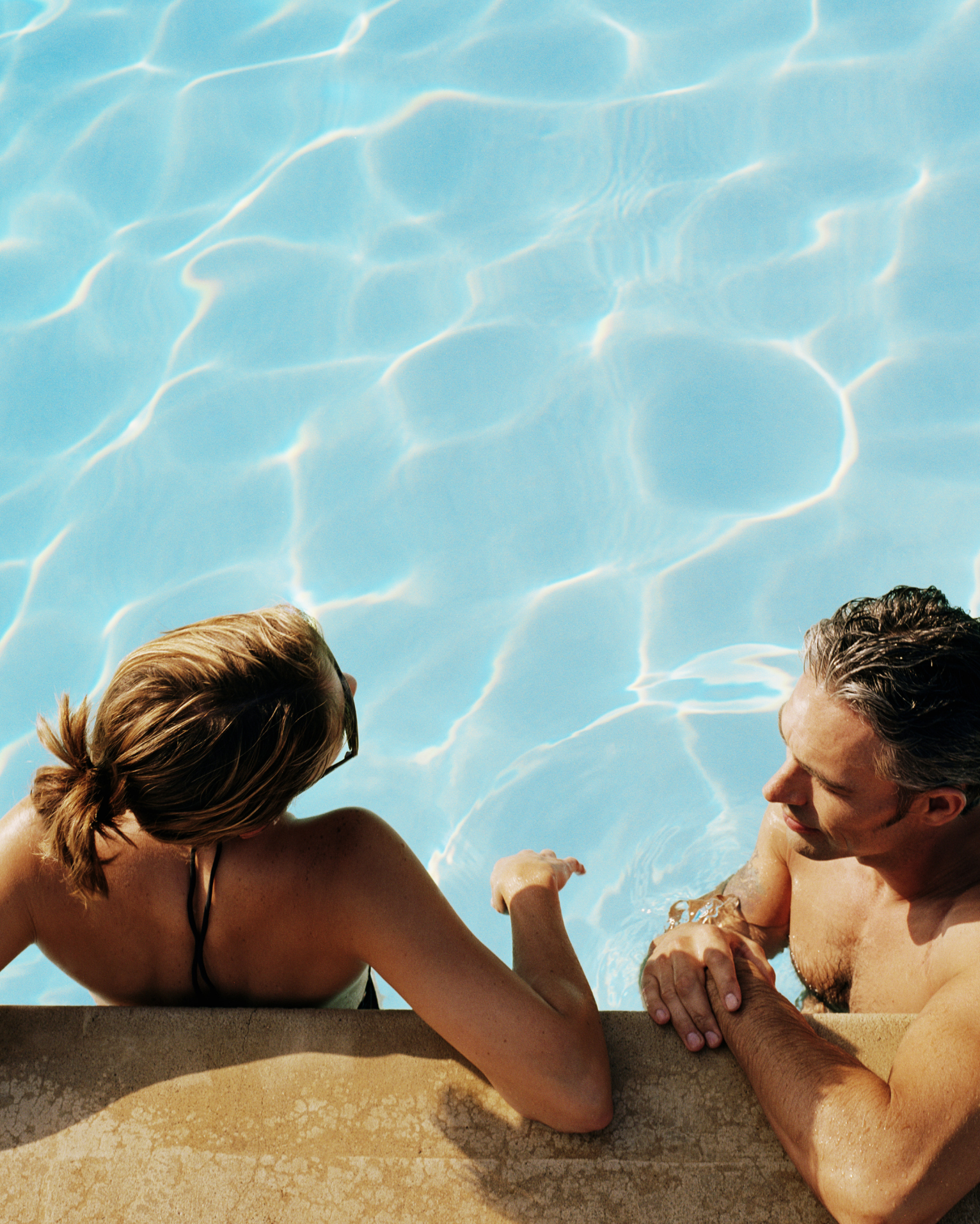 Two people relaxing at the edge of a swimming pool, leaning on the deck in bright blue water.