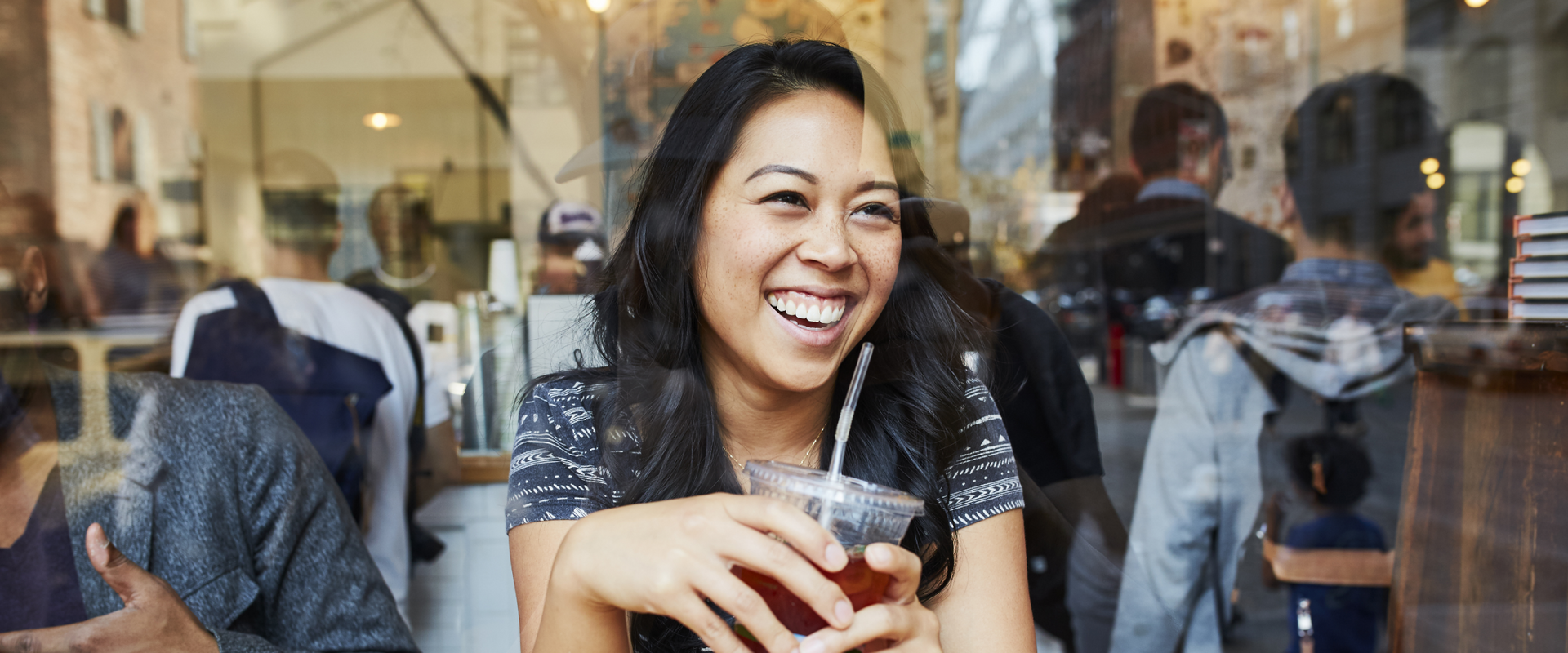 Smiling woman holding a drink in a cafe by a window, with people reflected in the glass