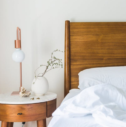 White bed with wooden headboard beside a round wooden nightstand and copper lamp in a bright bedroom