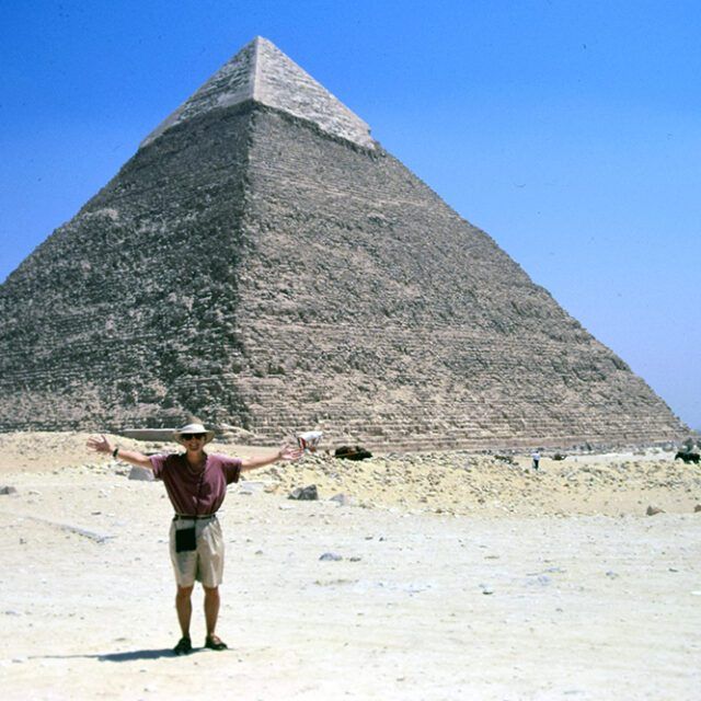 A man stands in front of a pyramid with his arms outstretched