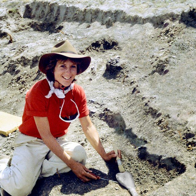 A woman wearing a cowboy hat is kneeling down in the dirt