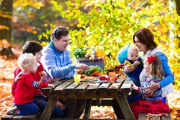 A family is sitting at a picnic table in the woods eating food.