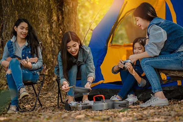 A group of young women are sitting around a campfire in front of a tent.