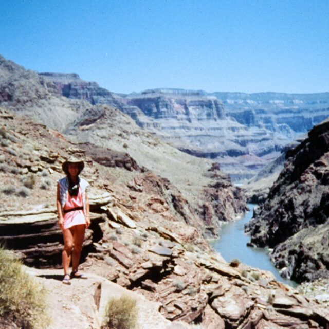 A woman in a cowboy hat stands on a rocky cliff overlooking a river