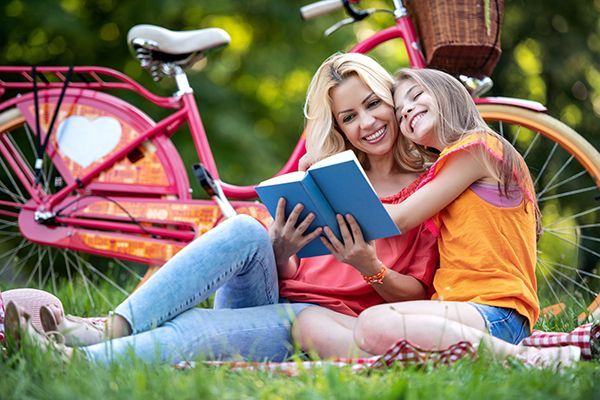 A mother and daughter are sitting on a blanket in the grass reading a book.