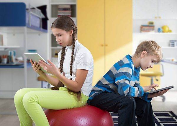A boy and a girl are sitting on an exercise ball looking at their tablets.