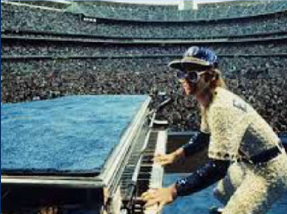 Elton John playing a piano in a baseball stadium, wearing a glittering outfit and sunglasses, with a large crowd in the background.