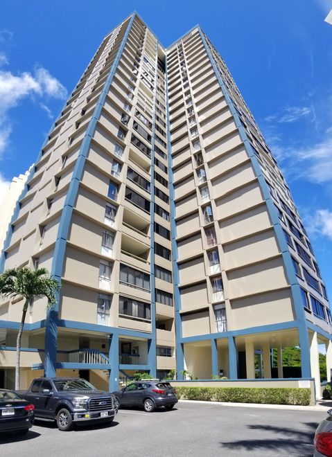 Two-story tan building with wraparound balcony and palm trees under a clear blue sky