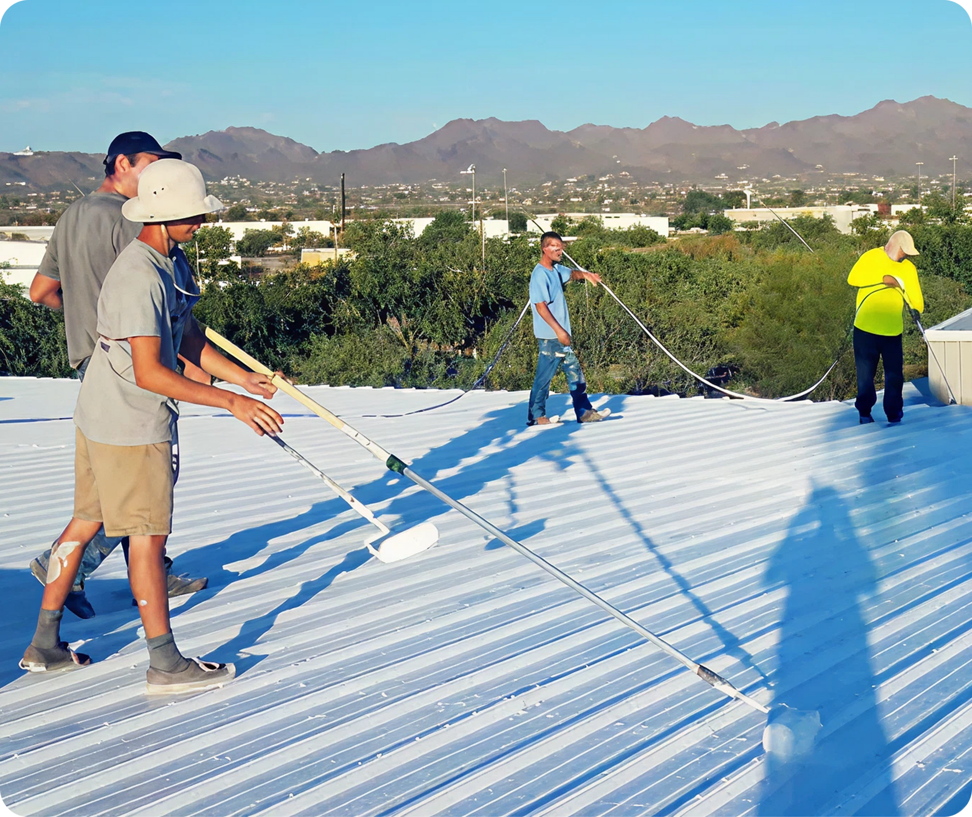 Workers painting a white corrugated roof under clear sky with mountains in the background